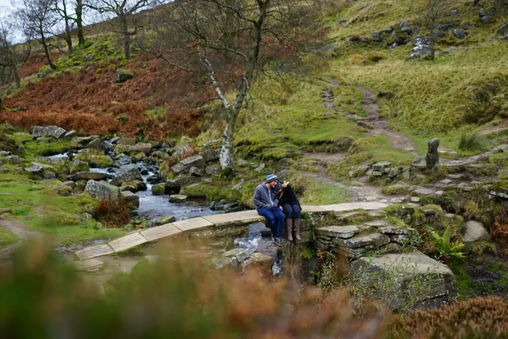 Couple sat on bridge with stream and green hills in Haworth