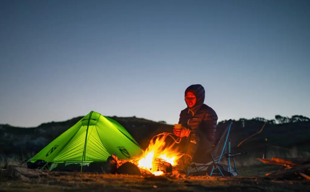 girl sat on Helinox Zero Chair beside fire and green tent at night