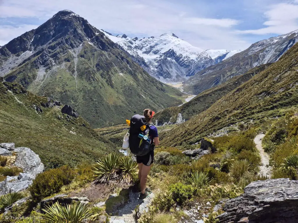 A girl walking the Rees Dart Track in New Zealand, with snow-capped mountains in front of her